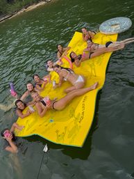 Group of smiling women lounging on a large yellow floating mat in a green lake near a tree-lined shore, holding colorful drinks and pool noodles on a sunny summer outing