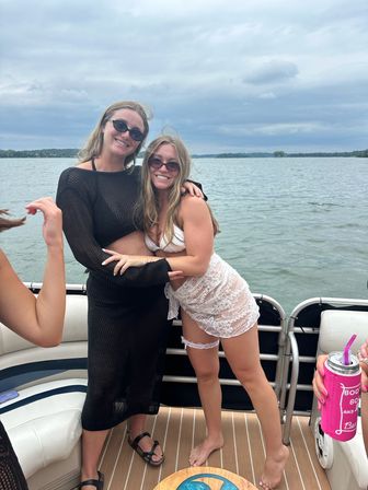 Two friends in sunglasses and summer outfits hugging on a pontoon boat with a lake and cloudy sky in the background, enjoying a casual summer boat day