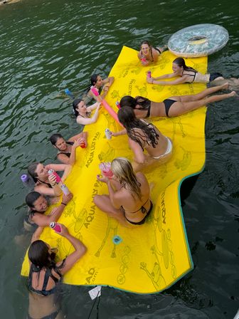 Group of friends in swimsuits lounging and laughing on a large yellow floating mat in a green lake, holding canned drinks, pool noodles and an inflatable tube nearby — outdoor summer lake float scene.