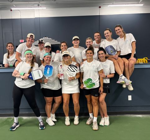 Indoor pickleball court team photo: a cheerful group of women in matching white shirts and athletic gear posing on a ledge, holding colorful paddles and a trophy after a match.