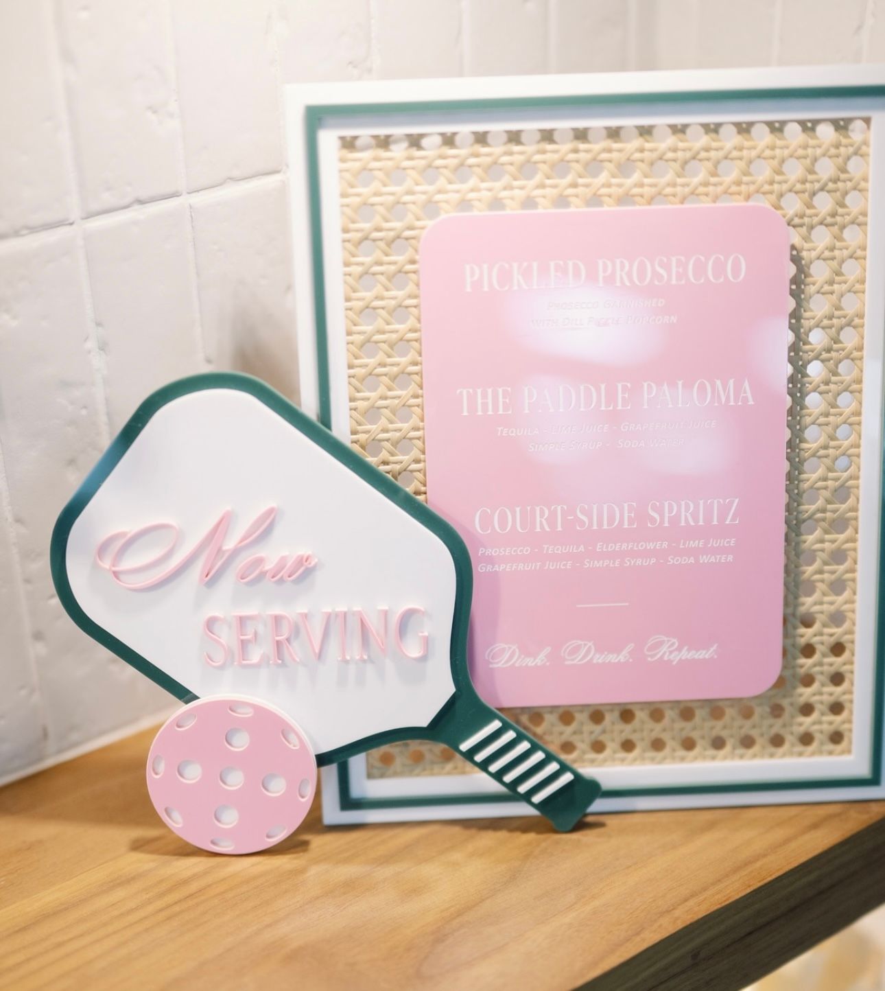 Pickleball-themed “Now Serving” paddle sign with a pastel pink ball beside a framed pink cocktail menu (Pickled Prosecco, Paddle Paloma, Court-Side Spritz) on a wooden countertop