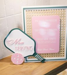 Pickleball-themed “Now Serving” paddle sign with a pastel pink ball beside a framed pink cocktail menu (Pickled Prosecco, Paddle Paloma, Court-Side Spritz) on a wooden countertop