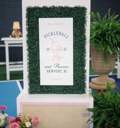 Decorative event sign reading Pickleball and Prosecco, Newport, RI, framed in faux boxwood on a blue pickleball court, flanked by wicker planter and fresh floral arrangements