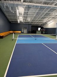 Indoor pickleball court with blue and light-blue courts, white net and lines, green perimeter, wooden bench seating along a dark wall, small bar area and exposed industrial ceiling in a recreation facility