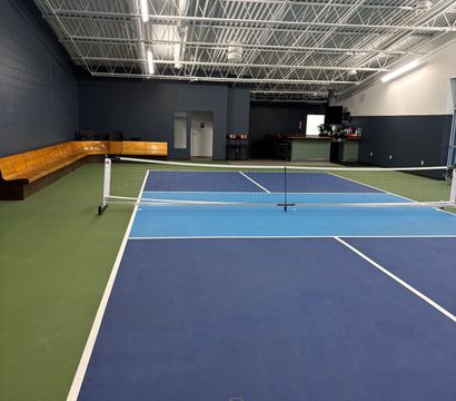 Indoor pickleball court with blue and light-blue courts, white net and lines, green perimeter, wooden bench seating along a dark wall, small bar area and exposed industrial ceiling in a recreation facility