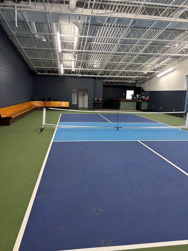 Indoor pickleball court with blue and light-blue courts, white net and lines, green perimeter, wooden bench seating along a dark wall, small bar area and exposed industrial ceiling in a recreation facility