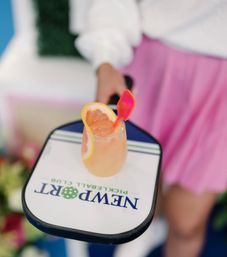 Hand holding a pickleball paddle with a bright grapefruit cocktail resting on it, garnished with a citrus slice and pink stirrer, blurred person in white top and pink skirt in the background.