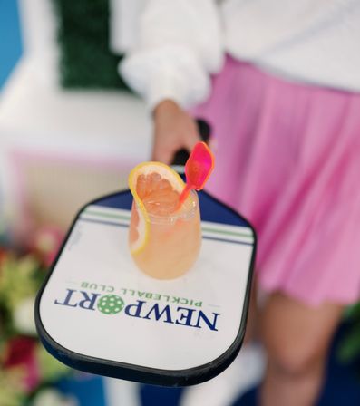 Hand holding a pickleball paddle with a bright grapefruit cocktail resting on it, garnished with a citrus slice and pink stirrer, blurred person in white top and pink skirt in the background.