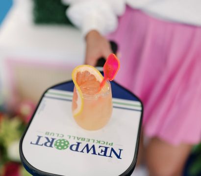 Hand holding a pickleball paddle with a bright grapefruit cocktail resting on it, garnished with a citrus slice and pink stirrer, blurred person in white top and pink skirt in the background.