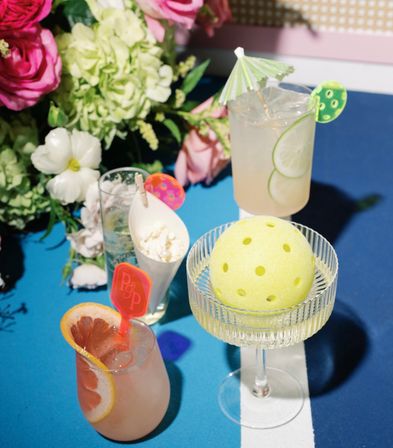Playful summer cocktail tablescape on a blue table: grapefruit- and lime-garnished drinks, a popcorn cone, floral centerpiece, and a yellow perforated decorative ball in a glass coupe.