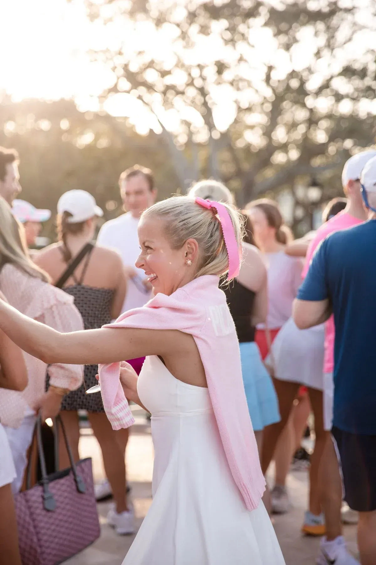 Smiling woman in a white dress with a pink sweater draped over her shoulders and matching ribbon reaching out at a sunlit outdoor gathering, surrounded by a casually dressed crowd.
