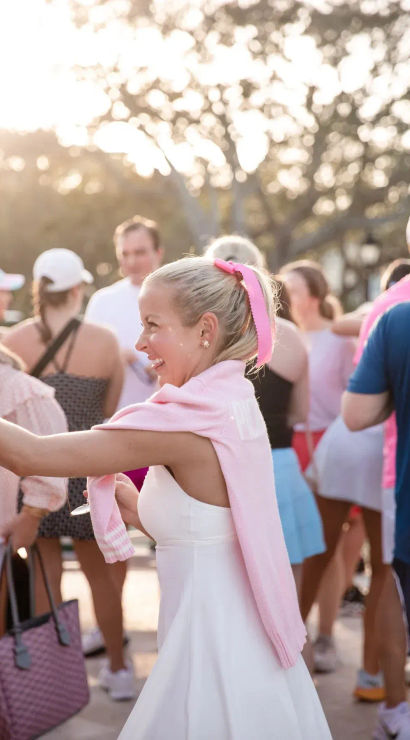 Smiling woman in a white dress with a pink sweater draped over her shoulders and matching ribbon reaching out at a sunlit outdoor gathering, surrounded by a casually dressed crowd.