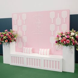 Cheerful pink event backdrop in Newport, RI with white pickleball paddle silhouettes, a striped bench with pillows, and tall bouquets of roses and mixed flowers.
