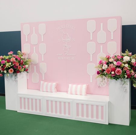 Cheerful pink event backdrop in Newport, RI with white pickleball paddle silhouettes, a striped bench with pillows, and tall bouquets of roses and mixed flowers.