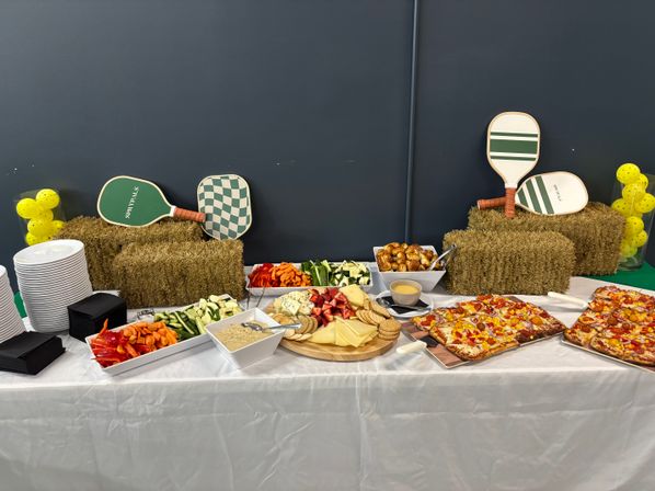 Indoor pickleball-themed catering spread with charcuterie board of cheeses, crackers and fruit, vegetable platters and dip, pretzel bites, rectangular pizzas, stacked plates and decorative paddles with yellow pickleball balls arranged on straw bales.