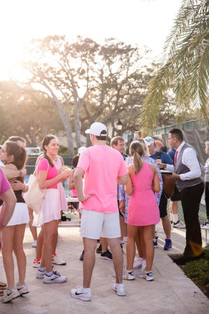 Sunlit outdoor tennis social at sunset — group of adults in pink sportswear chatting beside palm-lined courts on a tiled patio.