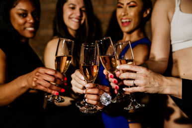 Close-up of a celebratory champagne toast — friends clinking sparkling wine flutes at a night out, colorful nail polish and rings, festive vibe.