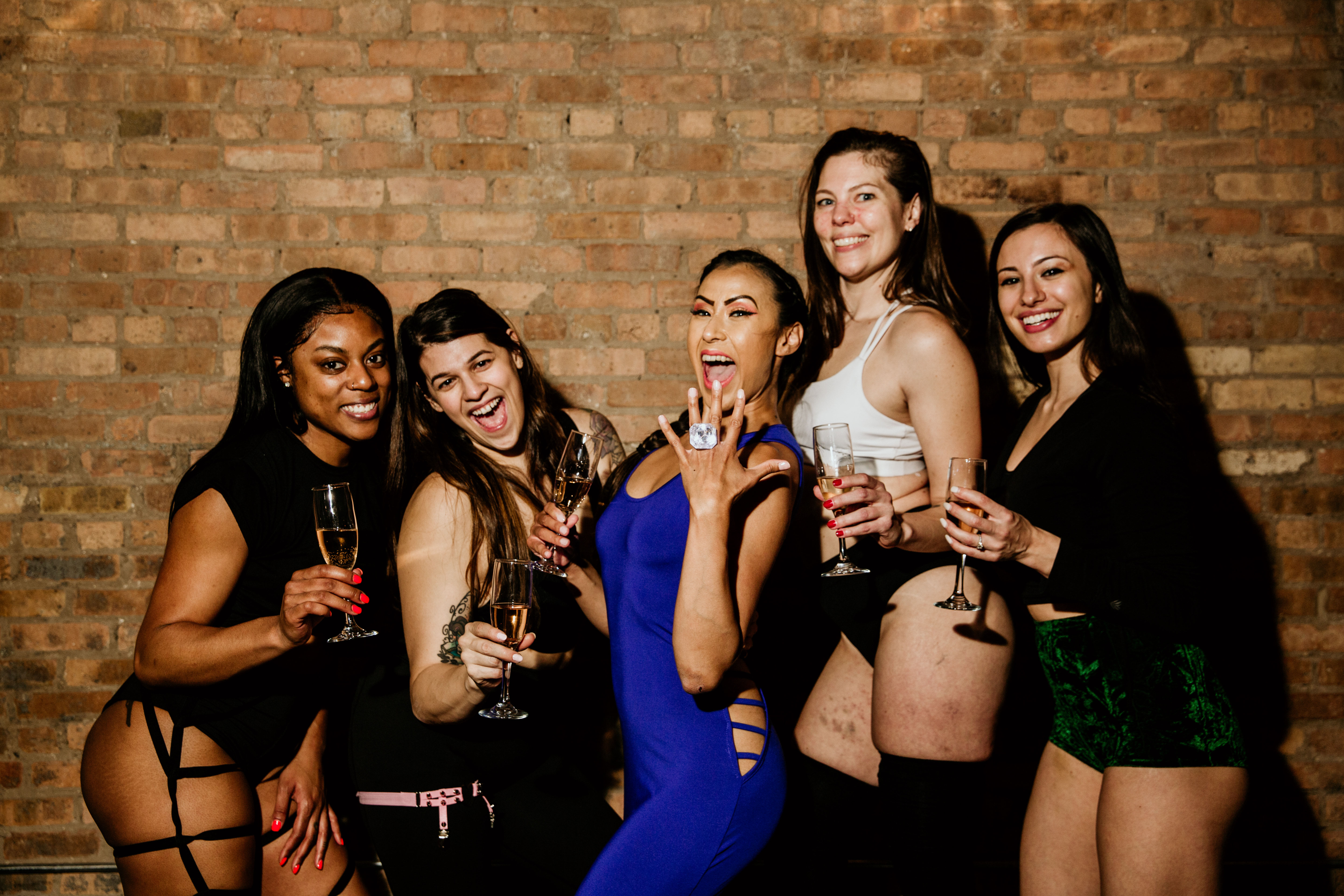 Five women in festive outfits holding champagne glasses and posing against an exposed brick wall, central woman in a blue dress proudly showing a ring—friends celebrating a night out or bachelorette party.
