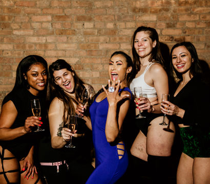 Five women in festive outfits holding champagne glasses and posing against an exposed brick wall, central woman in a blue dress proudly showing a ring—friends celebrating a night out or bachelorette party.
