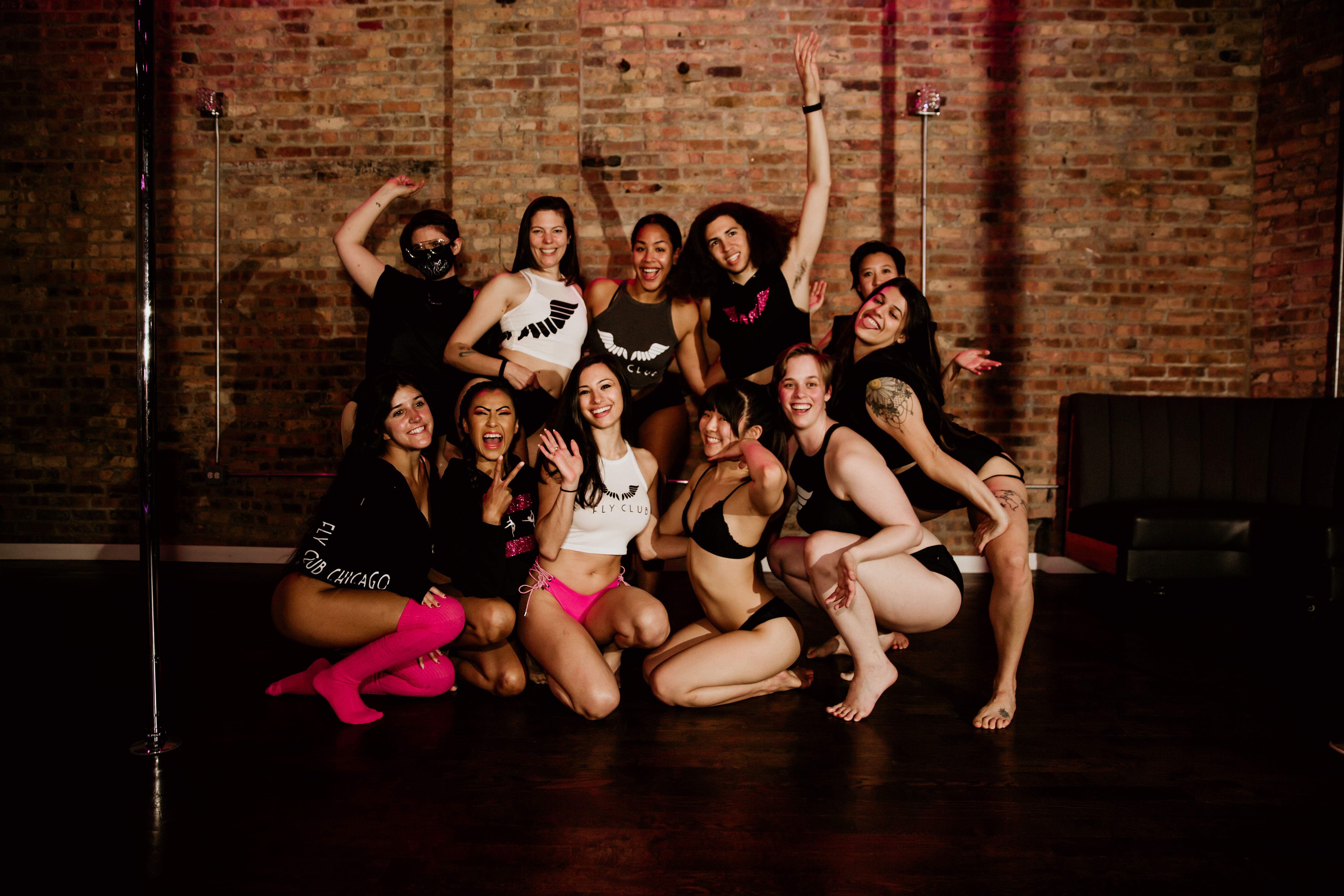 Smiling group of dancers in fitness and lingerie-style outfits posing together around a pole in a brick-walled urban dance studio.