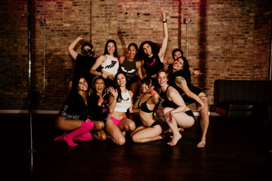 Smiling group of dancers in fitness and lingerie-style outfits posing together around a pole in a brick-walled urban dance studio.