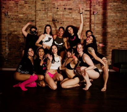Smiling group of dancers in fitness and lingerie-style outfits posing together around a pole in a brick-walled urban dance studio.