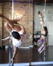 Pole fitness class in an urban studio with exposed brick and mirrors — students practicing pole climbs and spins in athletic wear and pink platform boots.