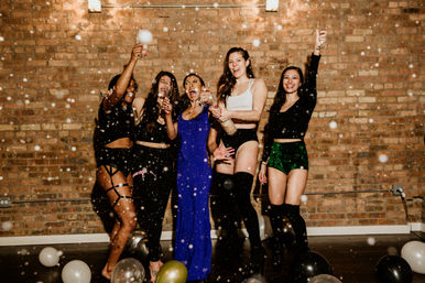 Five women celebrating in an urban loft with an exposed brick wall, popping champagne and cheering amid balloons and confetti