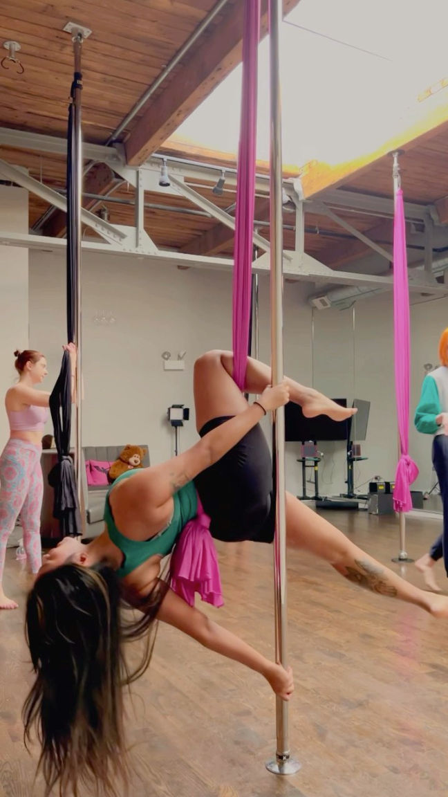 Pole fitness and aerial-silk class in a bright studio — performer hangs inverted from pink silk while gripping a chrome pole beneath a skylight and wooden beams on a hardwood floor.
