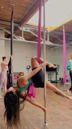 Pole fitness and aerial-silk class in a bright studio — performer hangs inverted from pink silk while gripping a chrome pole beneath a skylight and wooden beams on a hardwood floor.