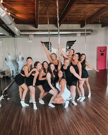 Group of smiling women in athletic wear posing around chrome dance poles in a bright pole-fitness studio with wood floors, mirrored wall, wing mural and industrial ceiling
