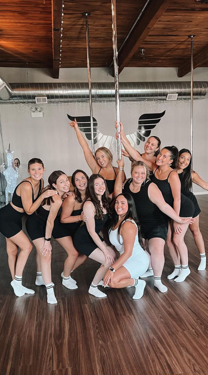 Group of smiling women in athletic wear posing around chrome dance poles in a bright pole-fitness studio with wood floors, mirrored wall, wing mural and industrial ceiling