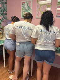 Three women standing in a pink boutique wearing matching The Bach Club t-shirts and summer denim shorts, browsing a display beneath tropical and sand-dollar wall art