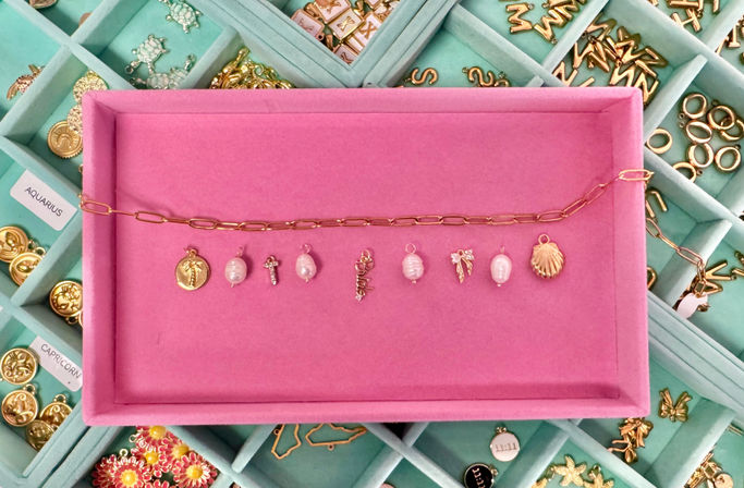 Pink felt jewelry tray with a gold chain and a row of small gold and pearl charms, displayed over a teal compartmented jewelry organizer with zodiac-labeled sections.
