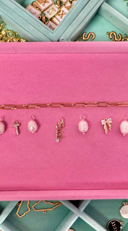 Pink felt jewelry tray with a gold chain and a row of small gold and pearl charms, displayed over a teal compartmented jewelry organizer with zodiac-labeled sections.