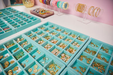 Turquoise compartment trays of gold letter charms, enamel pendants and small trinkets on a white counter with colorful bracelets displayed against a pink boutique wall