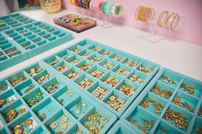 Turquoise compartment trays of gold letter charms, enamel pendants and small trinkets on a white counter with colorful bracelets displayed against a pink boutique wall