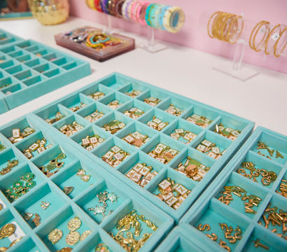 Turquoise compartment trays of gold letter charms, enamel pendants and small trinkets on a white counter with colorful bracelets displayed against a pink boutique wall