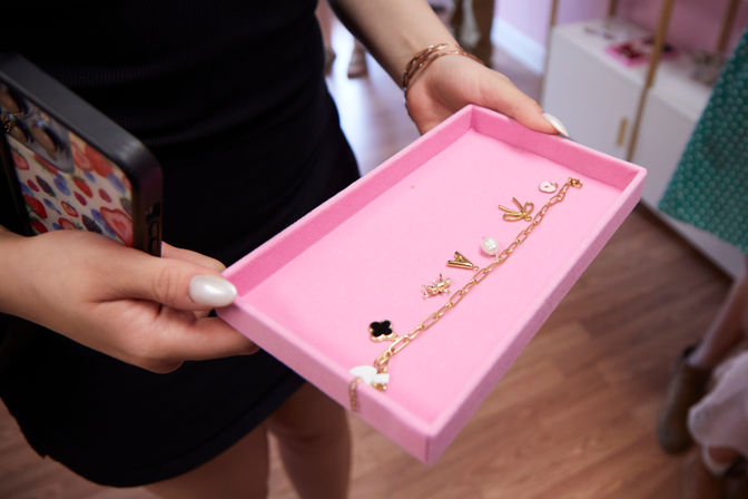 Hands holding a pink jewelry tray displaying a gold charm bracelet and small decorative charms, with a colorful-phone case visible — boutique jewelry display on hardwood floor.