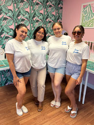 Four women in matching white "Team Bride" t-shirts smiling together for a bachelorette party inside a pink, tropical-print jewelry boutique with display trays and wood-look flooring.