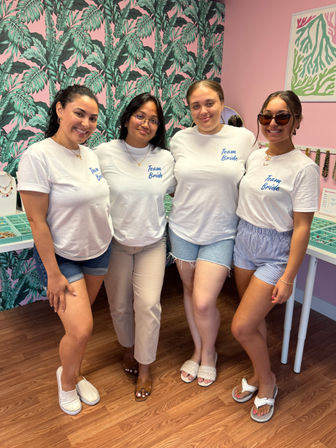 Four women in matching white "Team Bride" t-shirts smiling together for a bachelorette party inside a pink, tropical-print jewelry boutique with display trays and wood-look flooring.