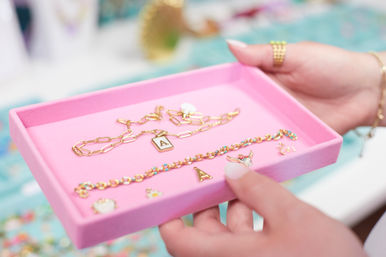 Close-up of hands holding a pink jewelry tray displaying a gold chain necklace with an 'A' initial charm, a colorful beaded bracelet and small enamel charms.