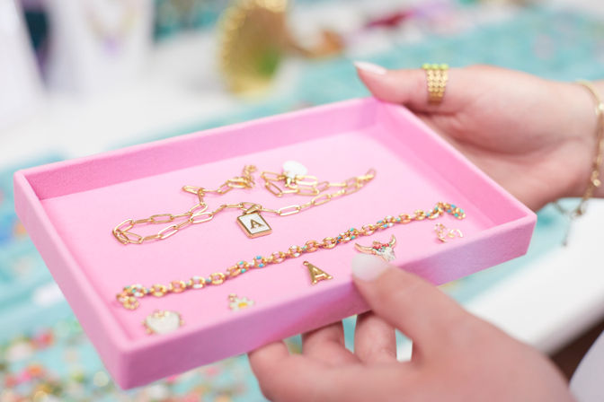 Close-up of hands holding a pink jewelry tray displaying a gold chain necklace with an 'A' initial charm, a colorful beaded bracelet and small enamel charms.
