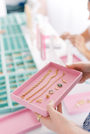 Hands holding a pink jewelry tray with gold chain bracelets and tiny enamel charms over a teal bead organizer on a bright craft table — handmade jewelry display.