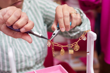 Close-up of hands using pliers to attach charms to a gold chain bracelet on a pink jewelry workstation in a craft studio, artisan handmade jewelry-making scene.