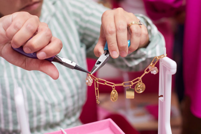Close-up of hands using pliers to attach charms to a gold chain bracelet on a pink jewelry workstation in a craft studio, artisan handmade jewelry-making scene.