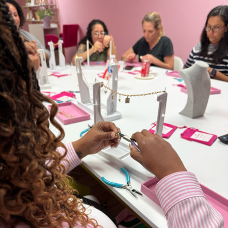 Close-up at a jewelry-making workshop in a pink craft studio: hands using pliers to attach gold charms to a bracelet chain on a white table surrounded by jewelry displays, tools, and pink trays.