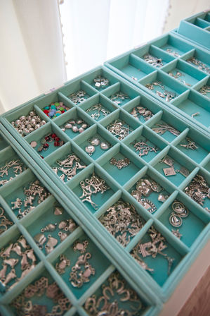 Close-up of a turquoise compartment jewelry organizer holding assorted silver charms, beads and pendants (hearts, shells, anchors) for jewelry-making and craft storage.