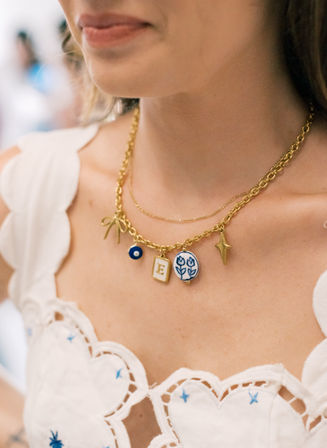 Close-up of a person’s neckline in a white embroidered sundress wearing layered gold charm necklaces with a blue enamel tulip charm, an 'E' initial pendant, evil-eye bead and star charm — playful summer jewelry detail.