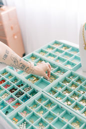 Tattooed hand reaching into mint-green compartment trays filled with gold charms, enamel pendants and colorful beads on a bright jewelry-making workstation
